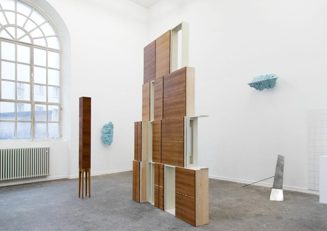 Foto: Ein Raum mit weißen Wänden, grauem, gefließten Steinboden und einem großen Fenster. An den Wänden befinden sich, auf unterschiedlicher Höhe, plastische Objekte. Im Raum stehen verschieden hohe Skulpturen auf unterschiedlichen Materialien.