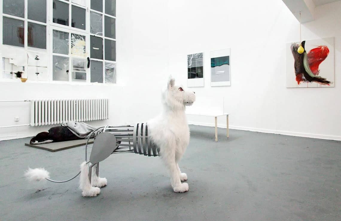 Foto: Ein Raum mit weißen Wänden, grauem Boden und einem großen Fenster. Teile des Fensters sind mit Folien, Tape und Papieren beklebt. An der Wand befinden sich mehrere flache, zum Teil gerahmte Objekte. Auf dem Boden stehen mehrere Skulpturen aus unterschiedlichen Materialien.