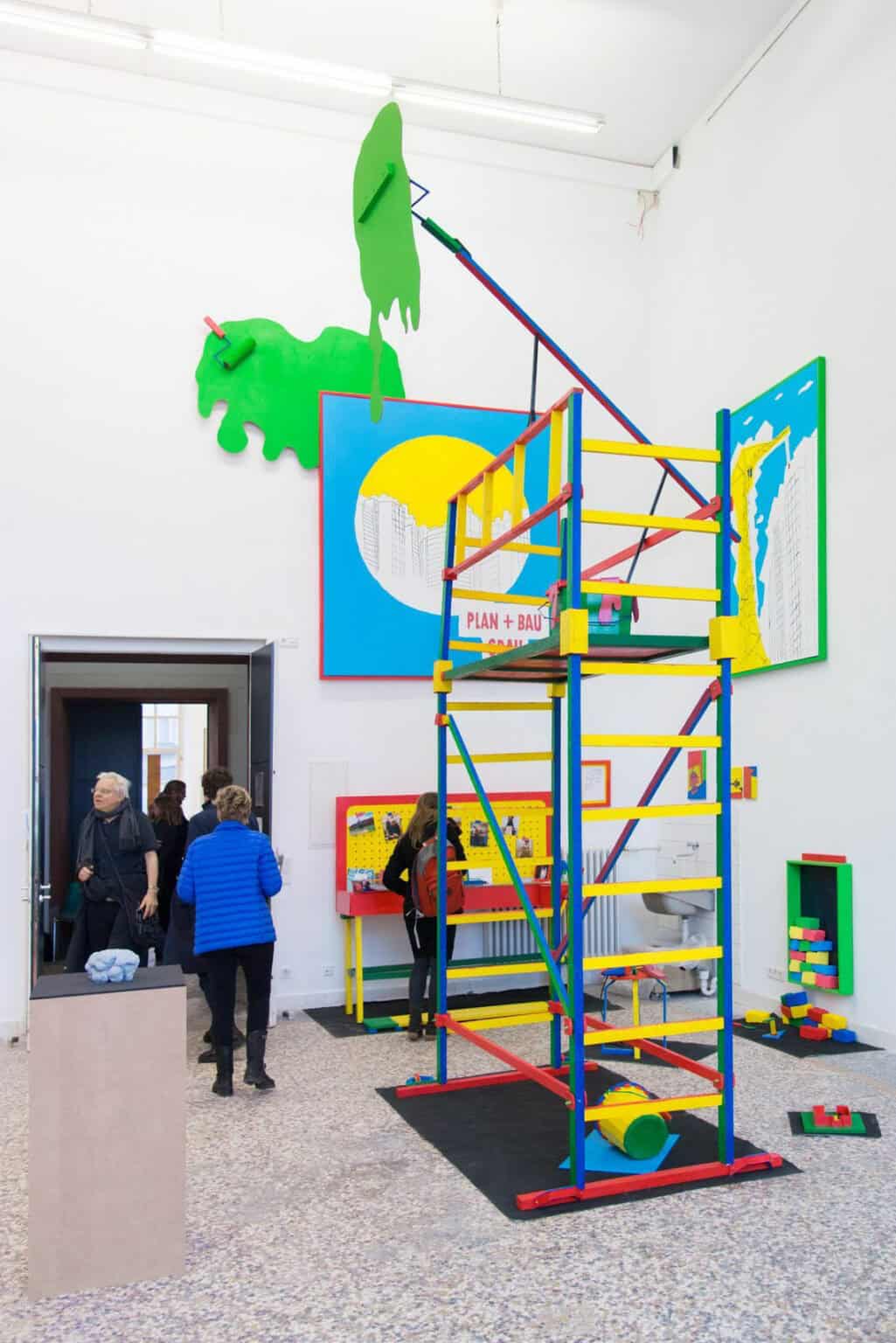 Foto: Ein Raum mit weißen Wänden, mehrfarbigem Steinboden und einer Tür. Im Raum befindet sich eine große, raumhohe Skulpturengruppe, die auch die Wände und den Boden bespielt. Im Raum befindet sich außerdem ein Sockel, auf dem eine kleine Skulptur liegt. Durch den Raum bewegen sich Menschen.