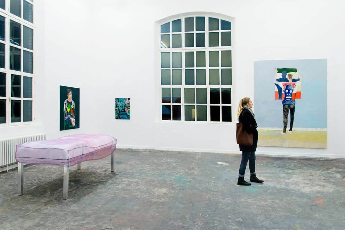 Foto: Ein Raum mit weißen Wänden, grauem Boden und zwei großen Fenstern. An den Wänden des Raumes befinden sich Leinwände in Mittel- und Großformaten. Im Raum befindet sich eine Skulptur und ein Mensch.