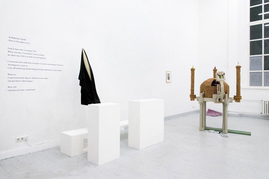 Foto: Ein Raum mit weißen Wänden, grauem Dielenboden und einem Fenster. Auf einer der Wände befindet sich schwarze Schrift und ein kleines, gerahmtes Objekt. Im Raum stehen Skulpturen und Sockel auf unterschiedlichen Materialien. Teile des Fensters sind mit halbtransparenten Papieren beklebt.