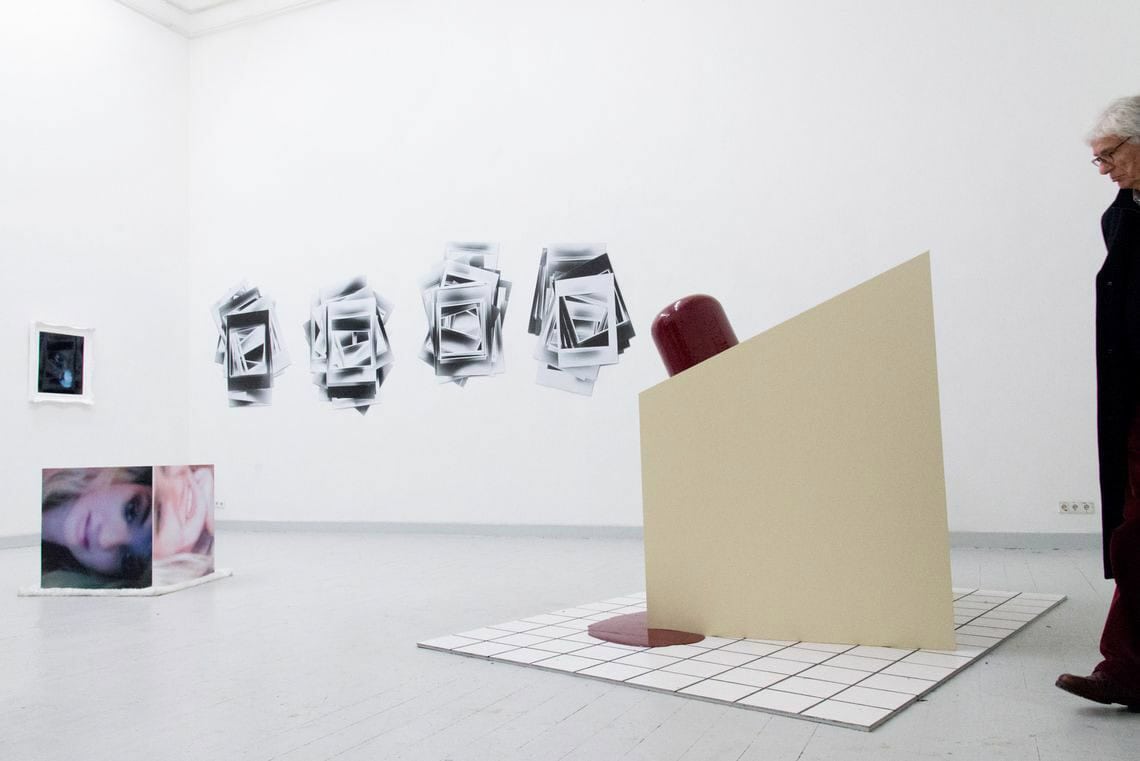 Foto: Ein Raum mit weißen Wänden und grauem Dielenboden. An der Wänden hängen mehrere flache Objekte auf ähnlicher Höhe. Auf dem Boden stehen zwei Skulpturen, die aus unterschiedlichen Materialien gefertigt sind. Im Raum befindet sich ein Mensch.