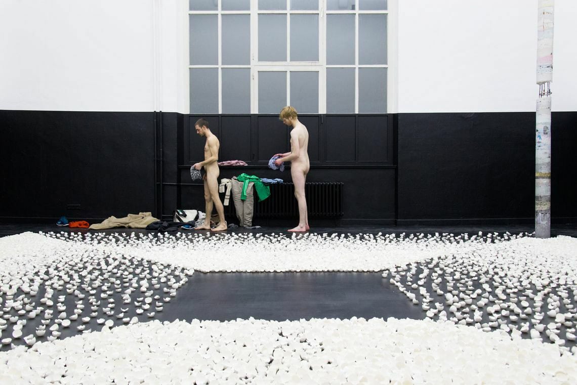 Foto: Ein Raum mit schwarzem Boden und einem Fenster. Der untere Teil er Wand ist schwarz gestrichen, der oberer Teil weiß. Die Fenster sind ebenfalls mit schwarze rund weißer Farbe bedeckt. Auf dem Boden des Raumes liegen viele kleine Objekte nah beieinander. Ein Quadrat in der Mitte des Raumes ist nicht von den Objekten bedeckt. hinten um Raum stehen zwei Nackte menschen, auf dem Boden liegt Kleidung. Rechts im Raum steht eine hohe, längliche Skulptur.