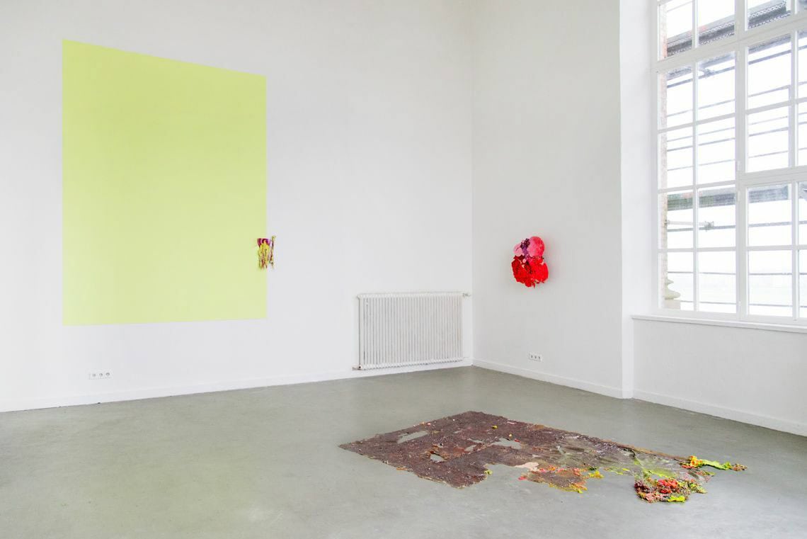 Foto: Ein Raum mit weißen Wänden, grauem Boden und einem großen Fenster. Auf die Wand ist ein gelbes Quadrat aufgemalt. Zwei kleine Objekte hängen auf der Wand. Auf dem Boden ist ein flaches, farbiges, krustiges Material ausgebreitet.