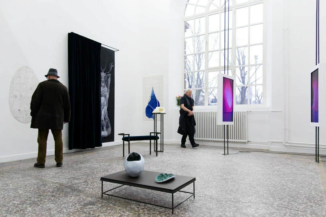 Foto: Ein Raum mit weißen Wänden, mehrfarbigem Steinboden und einem großen Fenster. An den Wänden des Raume befinden sich flache Objekte. Im Raum verteilt stehen mehrere Skulpturen auf dem Boden. Zwei Menschen bewegen sich durch den Raum.