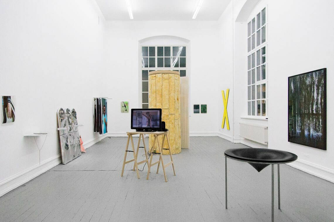 Foto: Ein Raum mit weißen Wänden, grauem Boden und großen Fenstern. An der Wand befinden sich mehrere flache und Plastische Objekte aus unterschiedlichen Materialien. Im Raum stehen mehrere Skulpturen und ein Bildschirm.