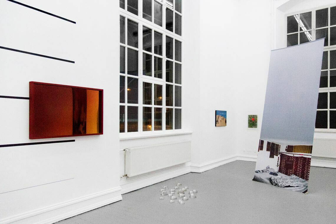 Foto: Ein Raum mit weißen Wänden, grauem Boden und großen Fenstern. An der Wand befinden sich mehrere flache Objekte. Eines von ihnen hängt auf schwarzen, auf die Wand aufgebrachten Streifen. Im Raum liegt eine Gruppe kleiner Objekte auf dem Boden. Von der Decke hängt ein bedruckte, hochformatiger Banner, der am Boden befestigt ist.