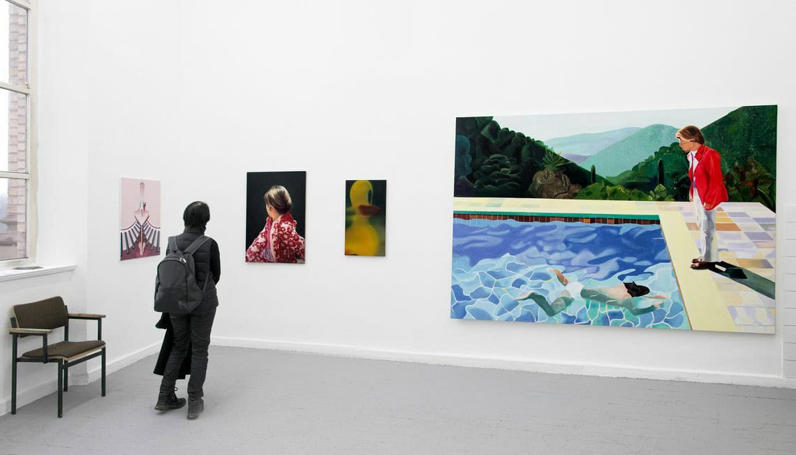 Foto: Ein Raum mit weißen Wänden und grauem Boden. An den Wänden hängen, auf ähnlicher Höhe nebeneinander, mehrere Leinwände in Mittel- und Großformaten. Im Raum befindet sich ein Mensch.