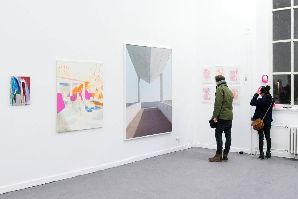 Foto: Ein Raum mit weißen Wänden, grauem Boden und einem großen Fenster. An der Wand beeiden sich mehrere flache Objekte. Manche von ihnen sind gerahmt. An einer Wand nahe des Fensters hängen zwei Kopfhörer. Im Raum befinden sich zwei Menschen.