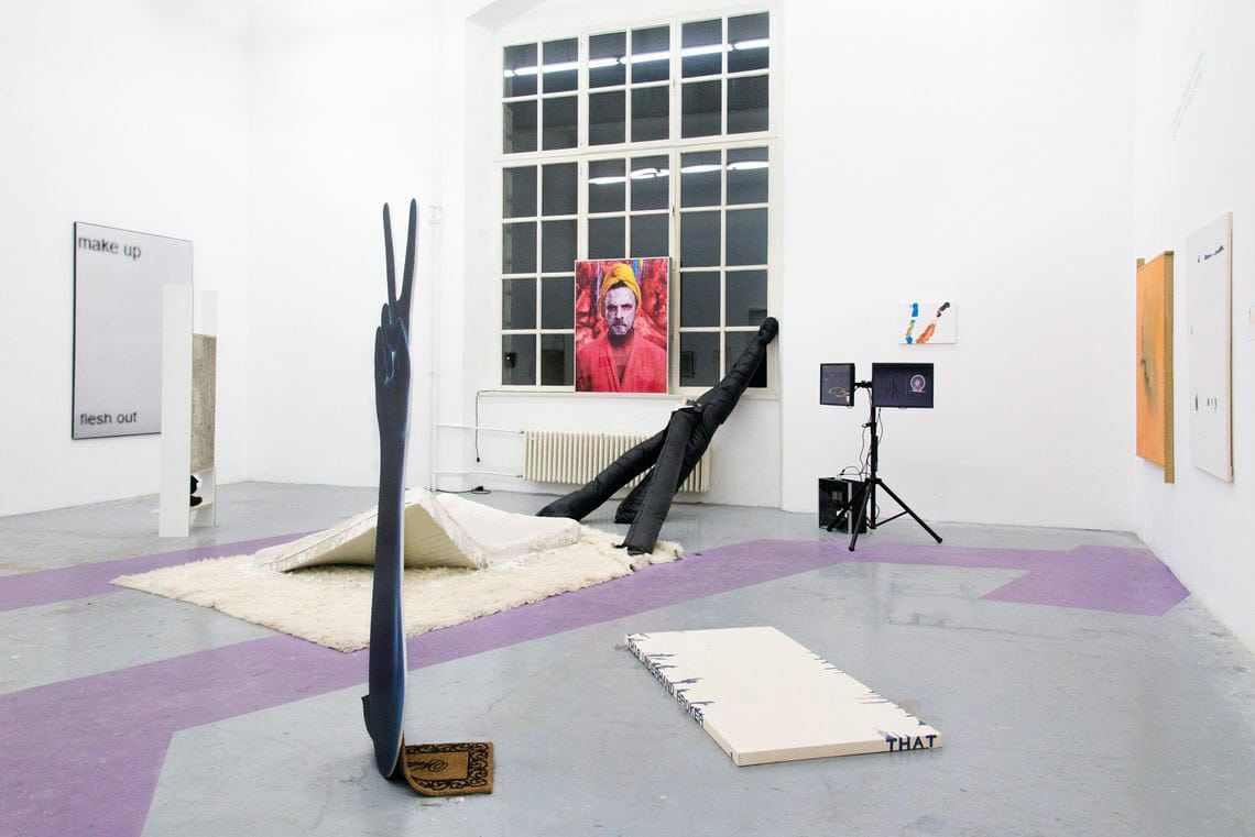 Foto: Ein Raum mit weißen Wänden, grauem Boden und einem großen Fenster. Teile des Bodens sind mit Lilianen, geometrischen Formen versehen. An den Wänden und an der Fensterbank hängen und lehnen flache Objekte. Im Raum stehen mehrere große Skulpturen und zwei Bildschirme, die sich auf einem Stativ befinden.