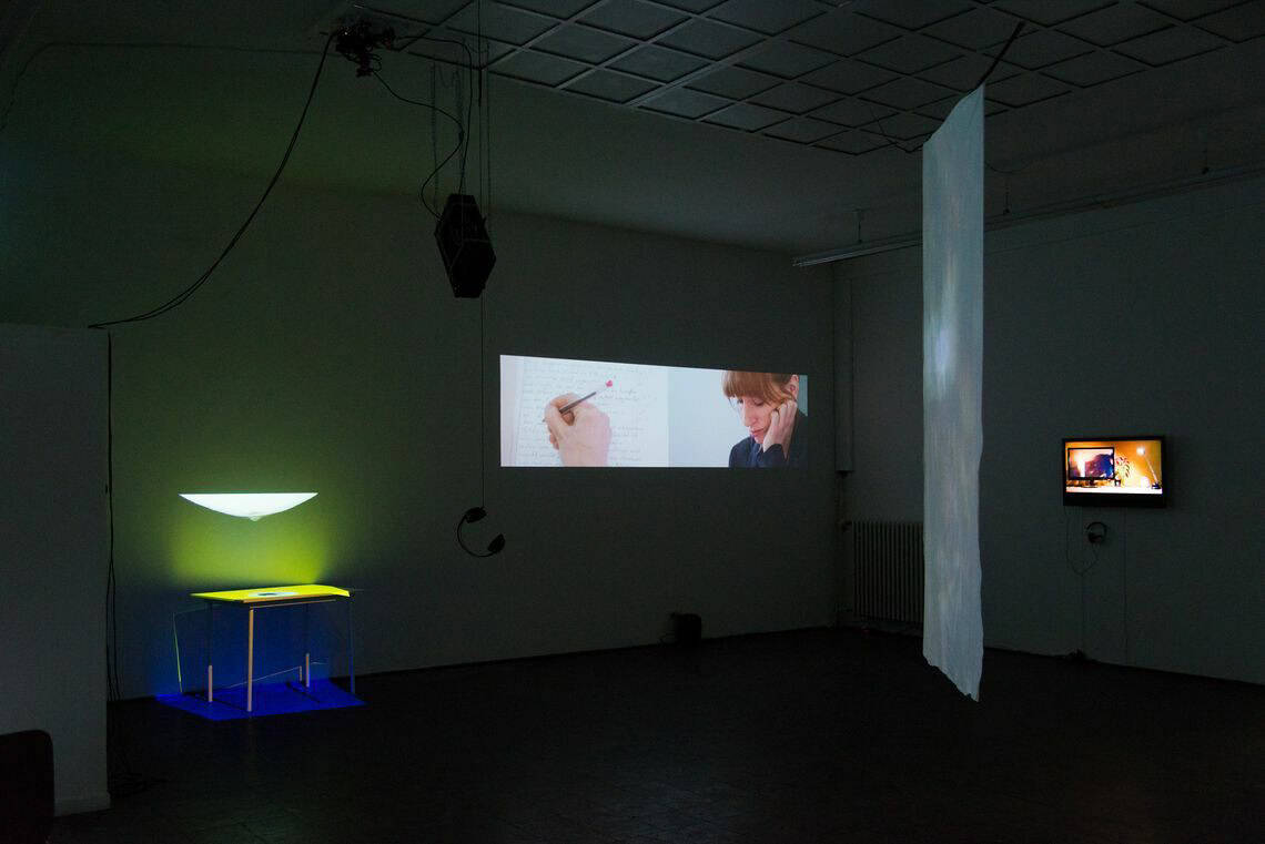 Foto: Ein dunkler Raum. An den Wänden befindet sich ein Bildschirm, eine Projektion und ein leuchtendes Objekt, unter dem eine Art Tisch an der Wand steht. Von der decke hängt ein ebenfalls leicht angeleuchtetes Material.