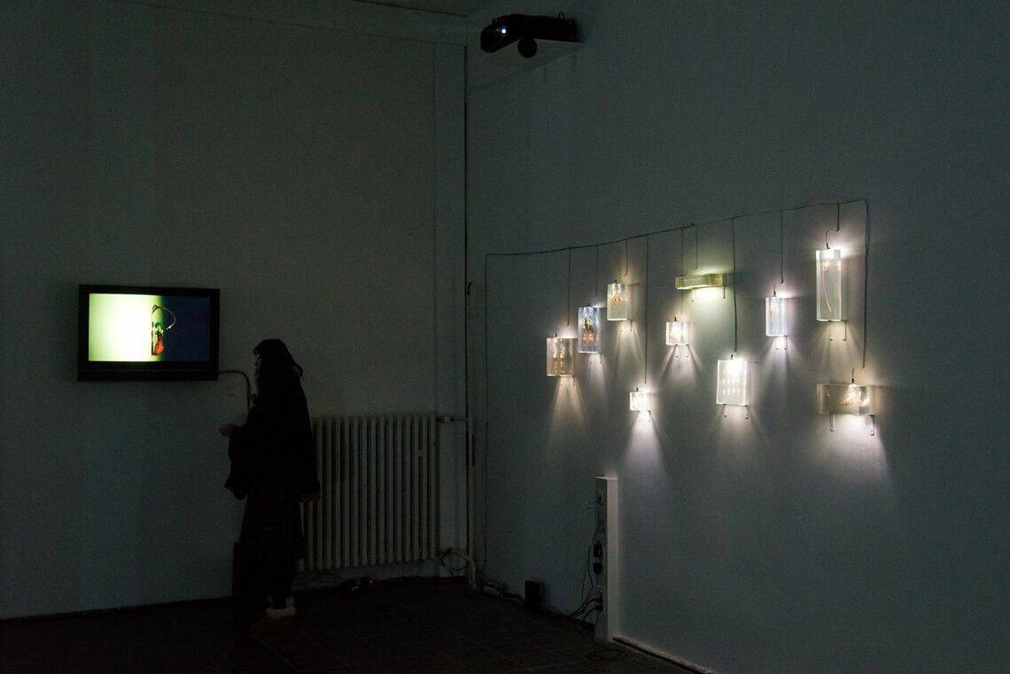Foto: Ein Dunkler Raum. An der Wand befindet sich ein Bildschirm und eine Konstruktion von an Seilen hängenden, leuchtenden Objekten.