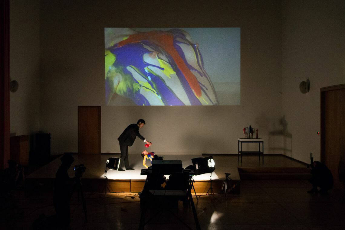 Foto: Ein Dunkler Raum. An die Wand wird eine große Projektion geworfen. Im Raum befindet sich Möbel, Skultpturen und Lichter sowie ein flächiges Podest. Auf dem Podest befinden sich zwei Menschen. Ein stehender Mensch gießt eine Flüssigkeit auf einen liegenden Menschen.