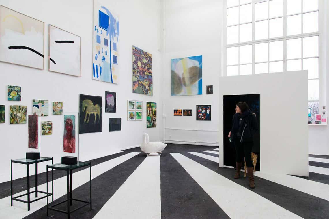 Foto: Ein Raum mit weißen Wänden und schwarz weißen, gestreiften Boden. An den Wänden hängen neben- und untereinander mehrere flache Objekte in Klein-, Mittel- und Großformaten. Manche von ihnen sind gerahmt. Auf dem Boden stehen Skulpturen. Durch den Raum bewegt sich ein Mensch.