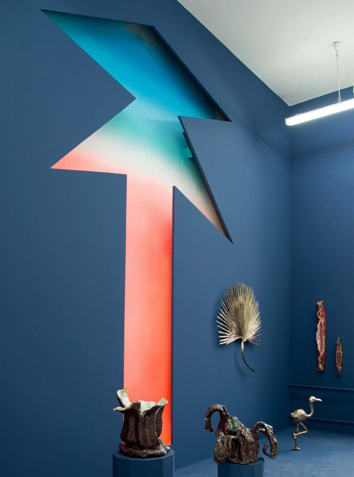 Foto: Ein Raum mit bleuen Wänden und blauem Boden. In einer der blauen Wände ist eine Aussparung. dahinter wird eine bunte Fläche sichtbar. An den Wänden hängen mehrere Objekte. Auch im Raum stehen Objekte, manche von ihnen befinden sich auf Sockeln.