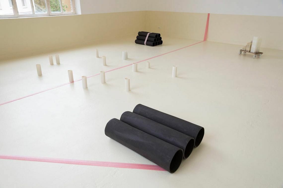 Foto: Ein Raum mit beigem Boden. Ein Streifen der Wand ist ebenfalls mit beige Farbe bemalt. Die darüberliegende Wand ist weiß. Über die beige Wand und den Boden verläuft rosanes Tape in einer Linie. Auf dem Boden liegen mehrere rohrartige Objekte in unterschiedlicher Ausrichtung und in unterschiedlicher Formation. Auf einem auf dem Boden befindlichen, kleinen Rollbrett liegen gestapelt weitere Gegenstände.