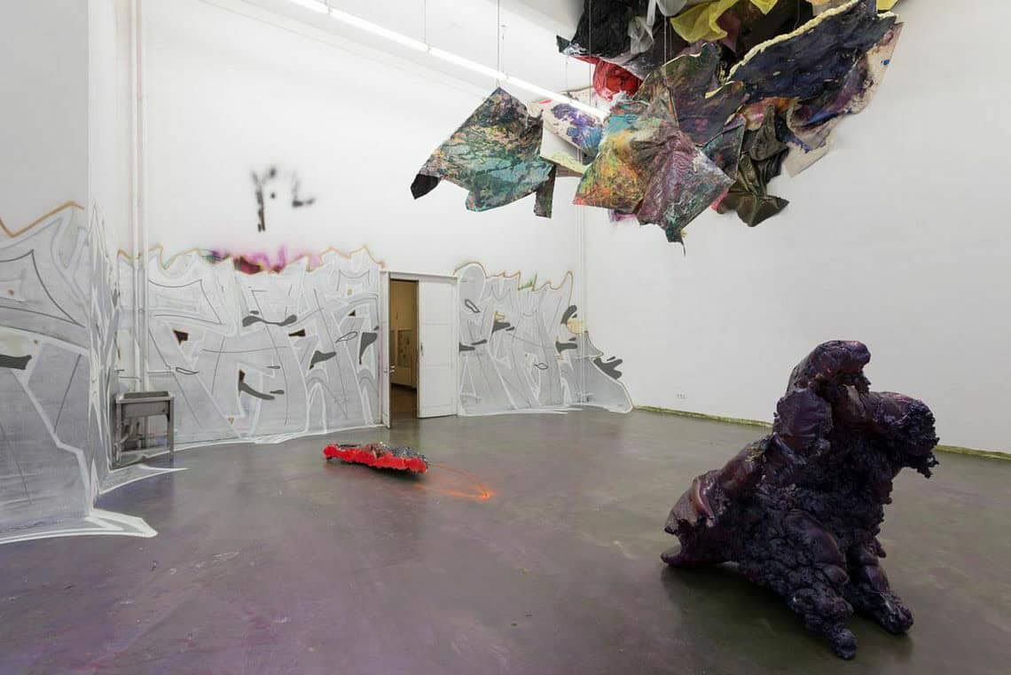 Foto: Ein Raum mit weißen Wänden und lila Boden. Große Teile der Wand sind mit Farbe besprüht. In einer Ecke des Raumes und Oben an der Ecke befindet sich eine Gruppe von Objekten. Im Raum stehen eine kleine und eine große Skulptur und ein orangefarbiger Fleck Sprühfarbe.