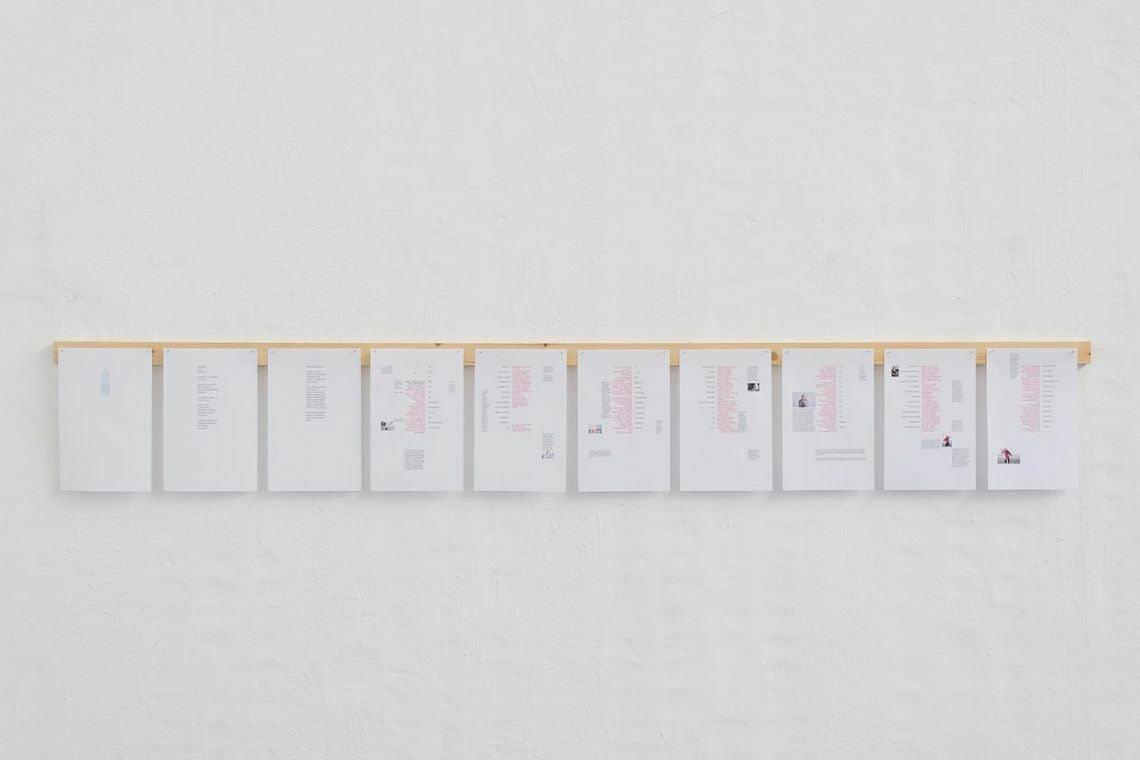 Foto: Eine weiße Wand, an der eine Holzlatte befestigt ist. An der Holzlatte hängen, in gleichen Abstand zueinander, mehrere A4 große, bedruckte Papiere.