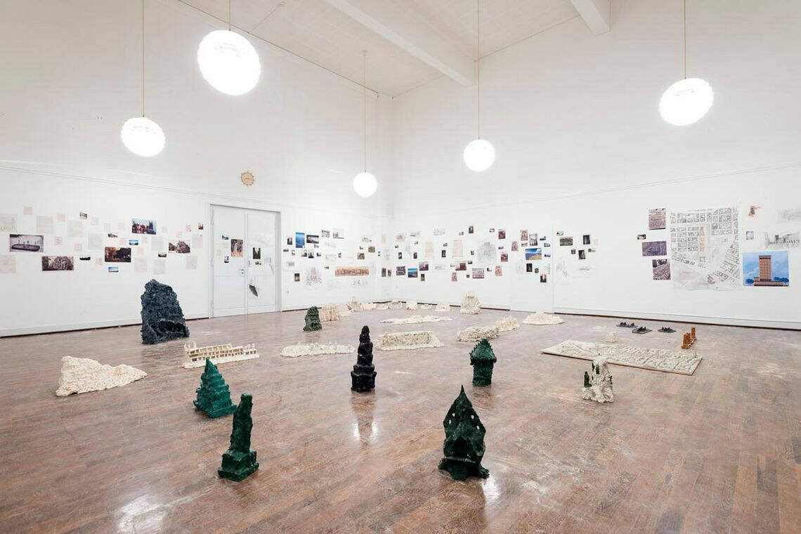 Foto: Ein Raum mit weißen Wänden und braunem Dielenboden. An den Wänden befindet sich eine große Menge an flachen Objekten und Papieren, die neben- und untereinander hängen. Im Raum verteilt auf dem Boden befinden sich ebenfalls viele Objekte. Stellenweise liegt Sand oder Staub auf dem Boden. Die Deckenbeleuchtung des Raumes spiegelt sich im Boden.