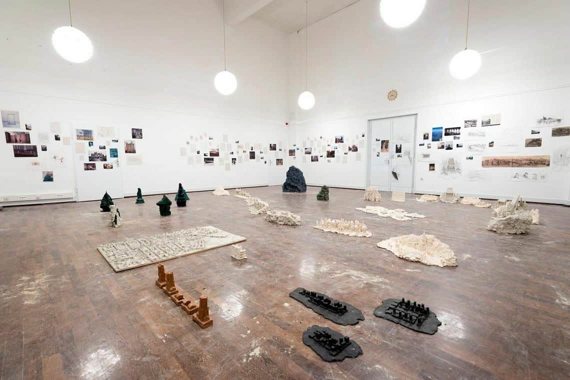 Foto: Ein Raum mit weißen Wänden und braunem Dielenboden. An den Wänden befindet sich eine große Menge an flachen Objekten und Papieren, die neben- und untereinander hängen. Im Raum verteilt auf dem Boden befinden sich ebenfalls viele Objekte. Stellenweise liegt Sand oder Staub auf dem Boden. Die Deckenbeleuchtung des Raumes spiegelt sich im Boden.