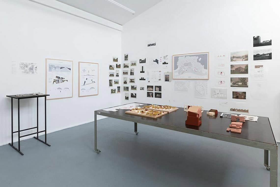 Foto: Ein Raum mit weißen Wänden und grauem Boden. An den Wänden befindet sich eine Vielzahl von gerahmten und ungerahmten Objekten. Im Raum steht ein Tisch, auf dem Objekte und Zettel liegen und eine Skulptur.