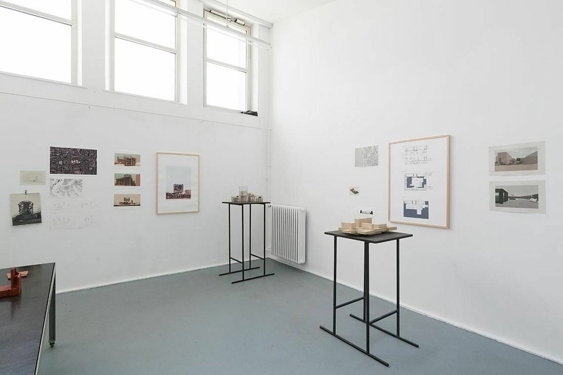 Foto: Ein Raum mit weißen Wänden, grauem Boden und einer hocheben im Raum sitzenden Reihe von Fenstern. An Den Wänden befinden sich gerahmte und ungerahmt Objekte. Im Raum stehen Tische, auf denen sich Objekte befinden und Skulpturen.