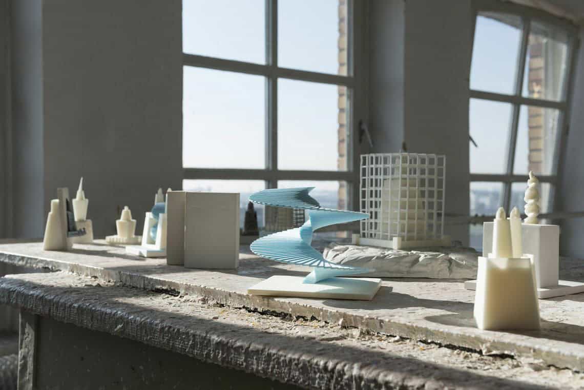 Foto: Ein Raum mit weißen Wänden, Fenstern und einem Tisch, der mit krustigem Material bedeckt ist. Auf dem Tisch steht eine Vielzahl modellartiger Objekte.
