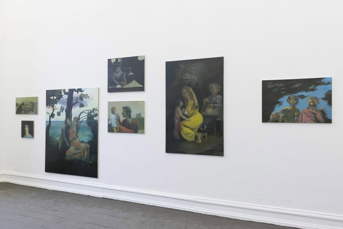 Foto: Eine weiße Wand und grauer Boden. An der Wand befinden sich, auf unterschiedlicher Höhe hängen, Leinwandarbeiten im Klein-, Mittel und Großformat.