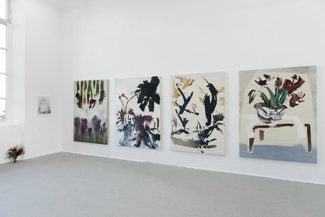 Foto: Ein Raum mit weißen Wänden und grauem Boden. An der Wand hängen vier großformatige Leinwandarbeiten, auf gleicher Höhe. An einer weiteren Wand hängt ein kleinformatiges Bild. Auf dem Boden steht ein Blumenstrauss.