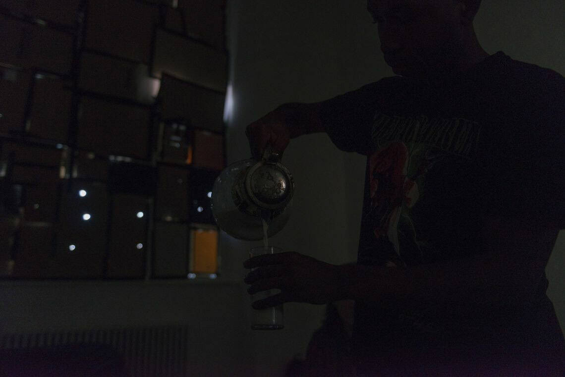 Foto: Ein dunkler Raum mit Fenster. Ein Mensch Schüttet Flüssigkeit aus einer Karaffe in ein Glas.