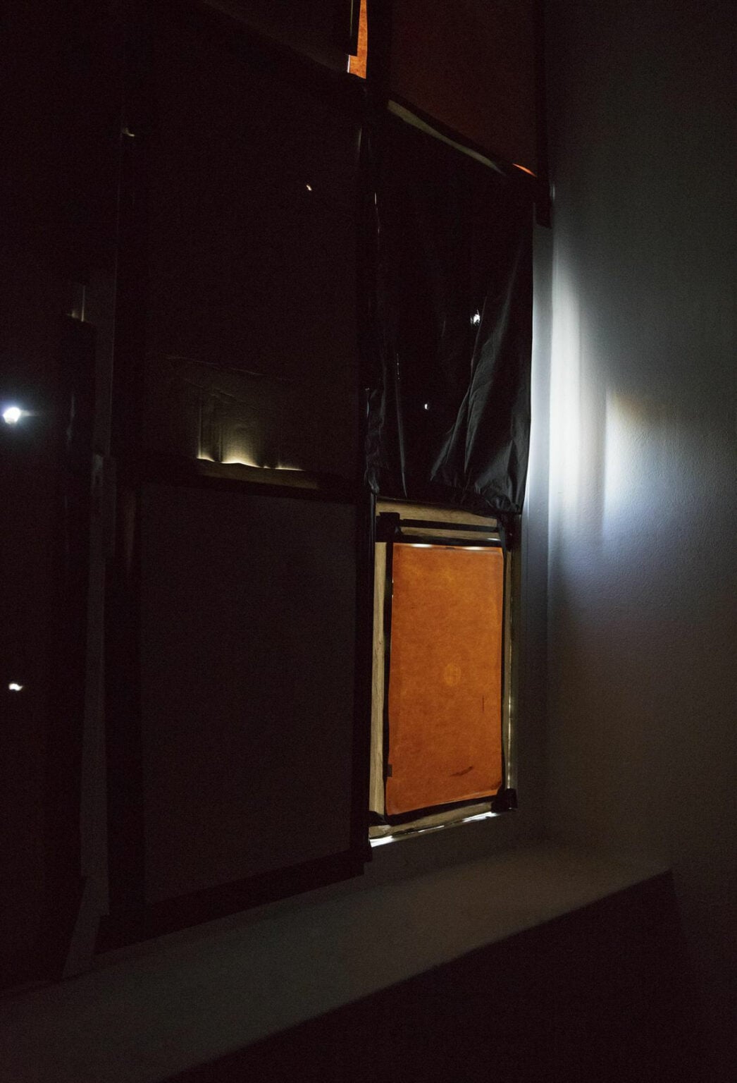 Foto: Detailaufnahme eines Fensters in einem dunklen Raum. Die Fensterscheiben sind mit unterschiedlichen Materialien bedeckt. Eines der Materialien ist lichtdurchlässig, es leuchtet etwas auf.