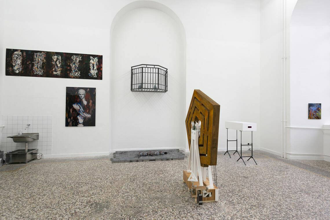 Foto: Ein Raum mit weißen Wänden, mehrfarbigem Steinboden und einer wandhohen, bogenförmigen Vertiefung. An der Wand des Raumes befinden sich mehrere Objekte auf unterschiedlicher Höhe. Im Raum stehen und liegen weitere Skulpturen auf dem Boden.