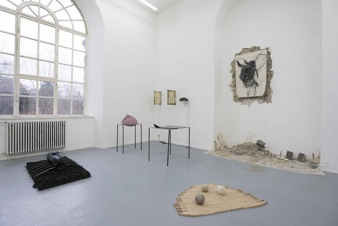 Foto: Ein Raum mit weißen Wänden, grauem Boden, einem großen Fenster und einer raumhohen, bogenförmigen Einbuchtung in einer der Wände. Innerhalb der Einbuchtung ist der Putzt stellenweise abgetragen. Der Schutt liegt unterhalb dieser Stelle auf dem Boden. Innerhalb dieser freigelegten Stelle hängt ein flaches, bemaltes Objekt. Daneben hängen weitere Objekte an der Wand. Im Raum stehen mehrere Skulpturen.