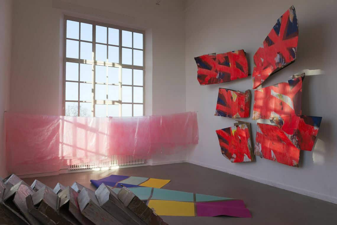 Foto: Ein Raum mit weißen Wänden, grauem Boden und einem großen Fenster. An der Wand des Raumes hängen mehrere große Objekte in einer Gruppe zusammen. Vor dem Fenster ist eine Schnur gespannt, an der halbtransparentes, rosanes Material hängt. Auf dem Boden liegen unterschiedliche Objekte.