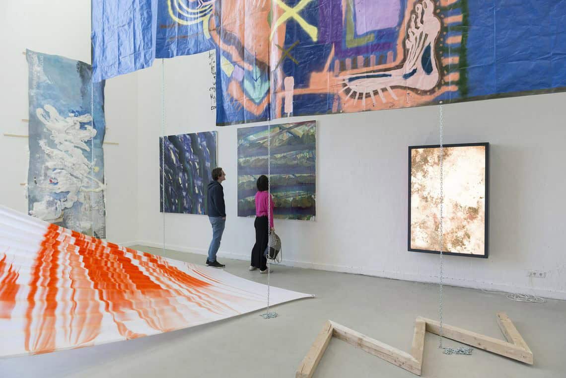 Foto: Ein Raum mit weißen Wänden und grauem Boden. An den Wänden des Raumes befinden sich mehrere großformatige Objekte aus unterschiedlichen Materialien. Auch von der Decke hängen flache, bemalte Materialien und Ketten. Auf dem Boden liegt eine zick-zack-förmige Skulptur. Zwischen dem Boden und einer der Wände ist ein farbiges Textil gespannt.