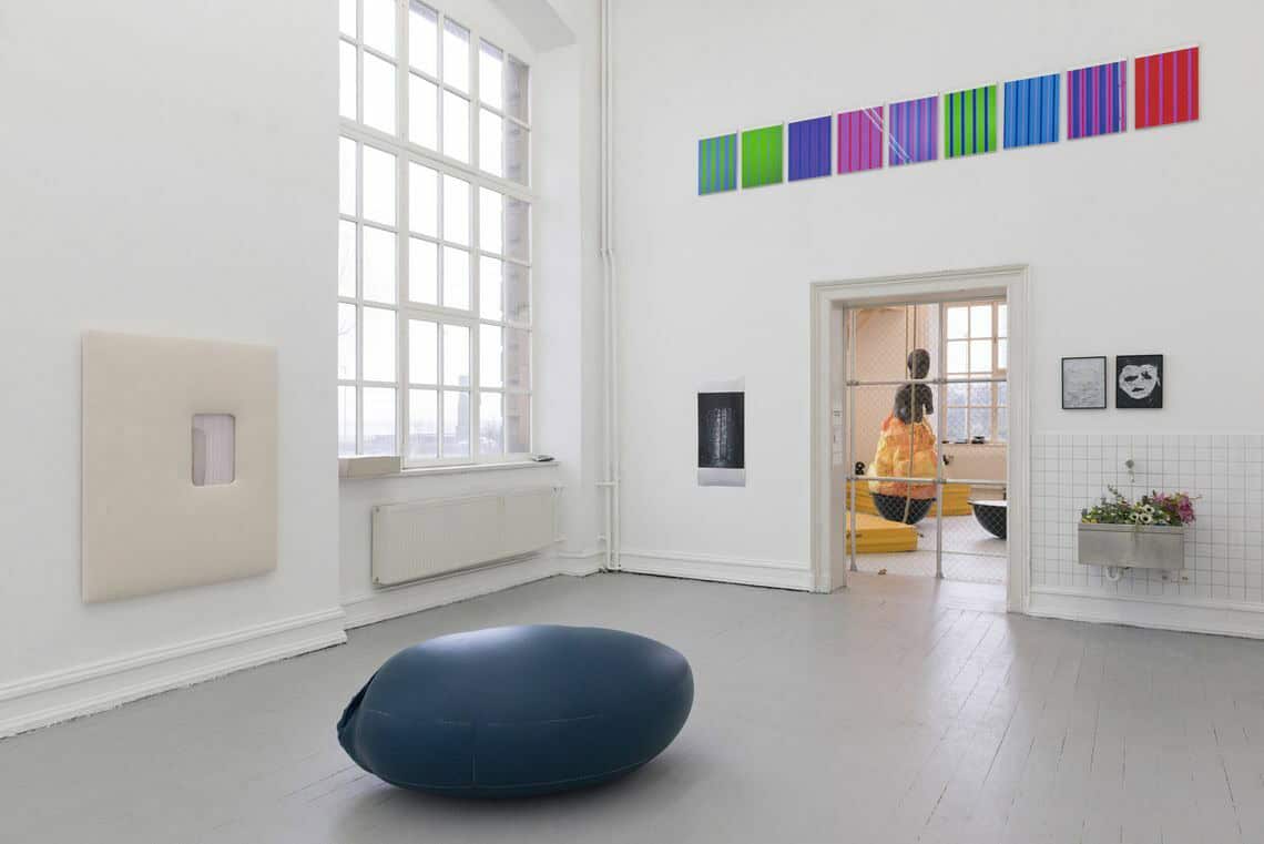 Foto: Ein Raum mit weißen Wänden, grauem Dielenboden und einem großen Fenster. An den Wänden des Raumes befinden sich, auf unterschiedlicher Höhe hängend, flache Objekte. Im Raum liegt ein weiteres Objekte. Durch den Raum hindurch sieht man in einen weiteren Raum, in dem sich eine Wand aus Gitter und mehrere große Objekte befinden.