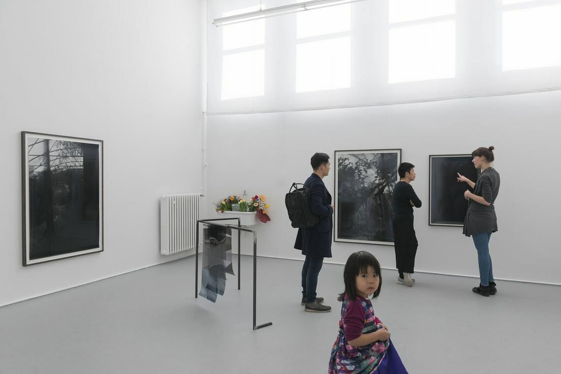 Foto: Ein Raum mit weißen Wänden und grauem Boden. Eine Reihe von Fenster befindet sich oben an einer der Wände. An der Wand hängen mehrere gerahmte Objekte. Im Raum steht eine Skulptur. Durch den Raum bewegen sich Menschen.