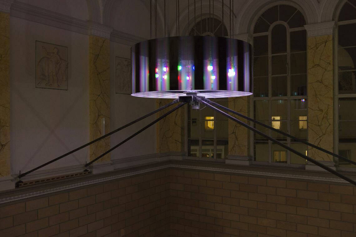 Foto: Ein hoher Raum mit drei großen Bogenfenstern. Von der Decke hängt ein kreisförmiges, transparentes, streifiges Objekt, in dessen Innern sich leuchtende Glühbirnen befinden. Das Objekt wird von vier Metallstreben gehalten, die an die Wand laufen.