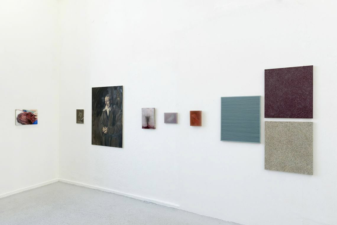 Foto: Ein Raum mit weißen Wänden und grauem Boden. An den Wänden hängen Leinwandarbeiten im Klein- und Mittelformat neben- und untereinander.