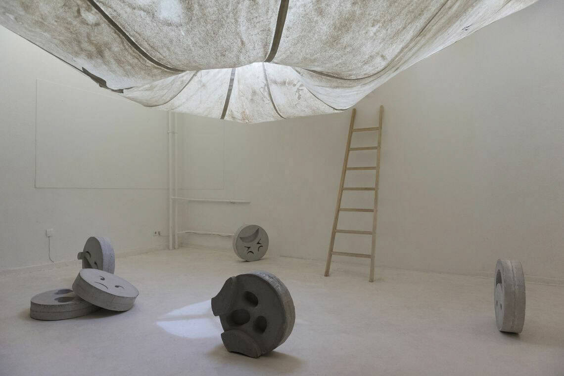 Foto: Ein Raum mit weißen Wänden und weißen Boden. Auf dem Boden des Raumes befinden sich mehrere runde Objekte, die stehen oder liegen. An der Wand lehnt eine Leiter. Von der Decke hängt ein Textil, dass in seiner Mitte trichterartig nach innen zusammenläuft.