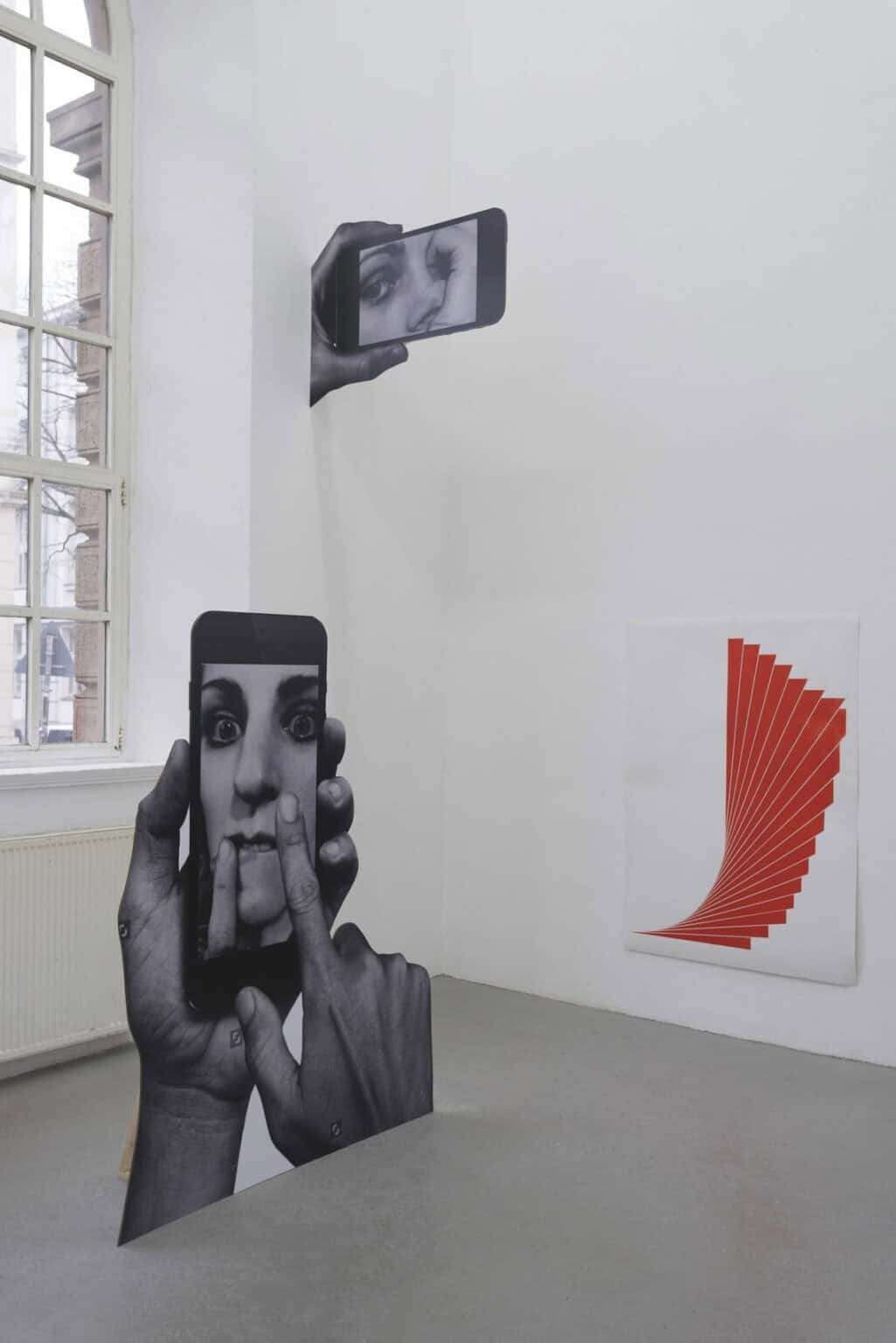 Foto: Ein Raum mit weißen Wänden, grauem Boden und einem Fenster. An den Wänden hängen zwei Objekte, eines von ihnen flach an der Wand, das andere steht von der Wand Ab. Auf dem Boden steht ebenfalls ein Objekt, das einer Art Papieraufsteller gleicht.