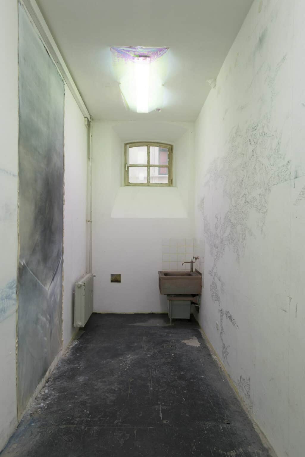Foto: Ein schmaler Raum mit kleinem, hochgelegenen Fenster und grellem Deckenlicht. An einer der Wände hängt eine mit Farbe bemalte, umaufgespannte Leinwand. Die gegenüberliegende Wand ist mit deinen grauen Elementen bemalt. Im Raum befindet sich ein Waschbecken. Daneben hängt ein kleinformatiges Objekt an der Wand. Der Boden ist staubig.
