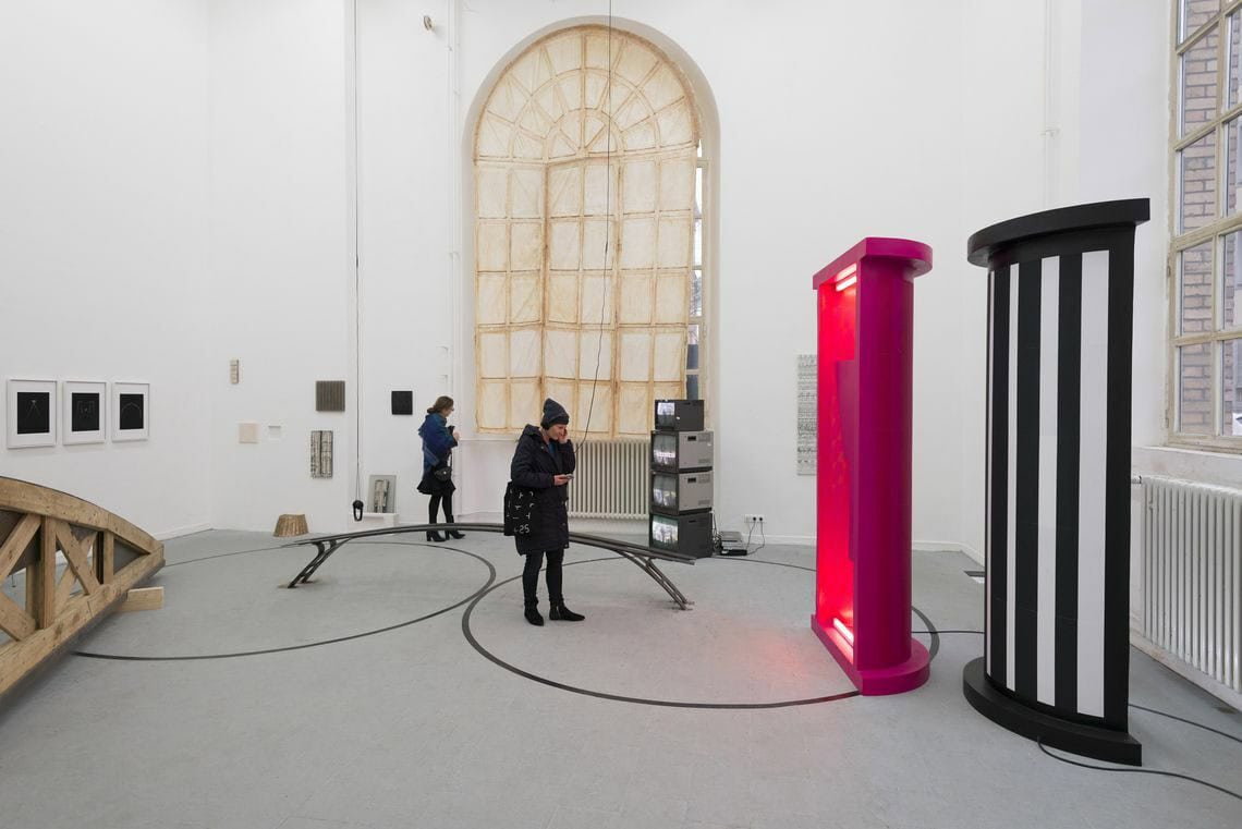 Foto: Ein Raum mit weißen Wänden, grauem Boden und zwei großen Bogenfenstern. Auf den Boden sind zwei bleichgroße, schwarze Kreise aufgemalt, die sich berühren. An den Wänden hängen viele mittel- und kleinformatige Objekte auf unetrschiedlciehr Höhe. Im Raum stehen mehrere große Skulpturen. Eine der Skulpturen liegt mit ihren beiden Enden jeweils in einem der aufgemalten Kreise auf dem Boden auf. Eines der Fenster ist mit einem bemalten Textil bedeckt, dass die gleiche Form hat wie das Fenster.