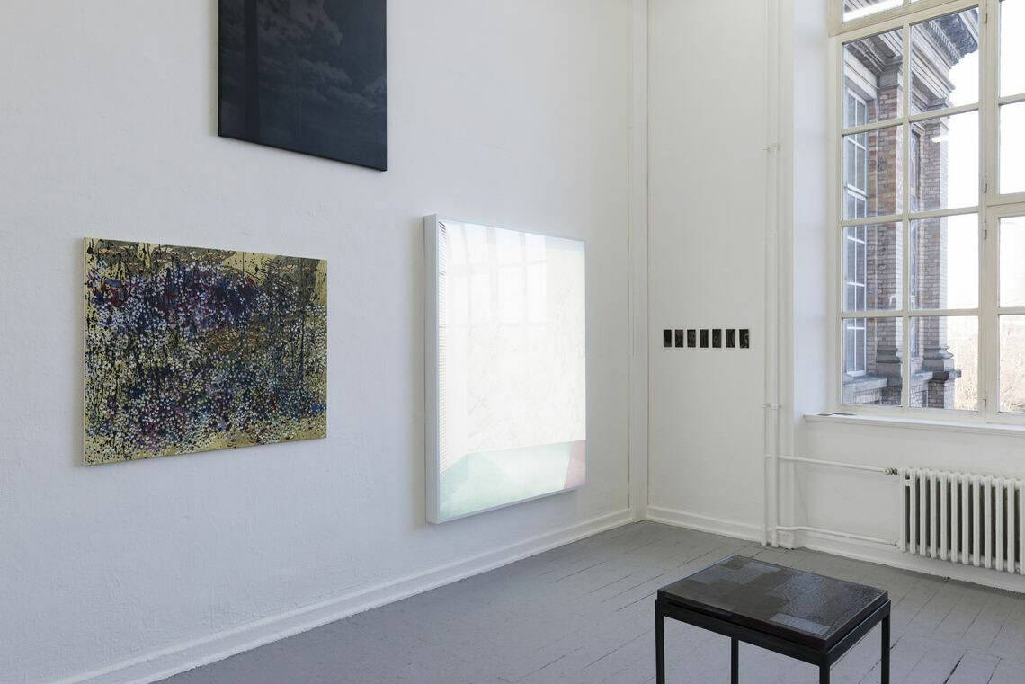 Foto: Ein Raum mit weißen Wänden, grauem Dielenboden und Fenster. An den Wänden hängen, auf unterschiedlicher Höhe, mehrere flache Objekte. Im Raum steht eine Skulptur. An einer weitern Wand hängen kleinformatige Objekte in einer Reihe nebeneinander.