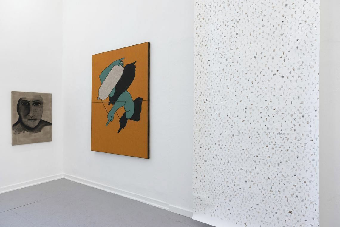 Foto: Ein Raum mit weißen Wänden und grauem Boden. An der Wand hängen zwei flache Objekte im Klein- und Mittelformat. Von der Decke hängt eine bemalt Rolle, die kurz über dem Boden endet.