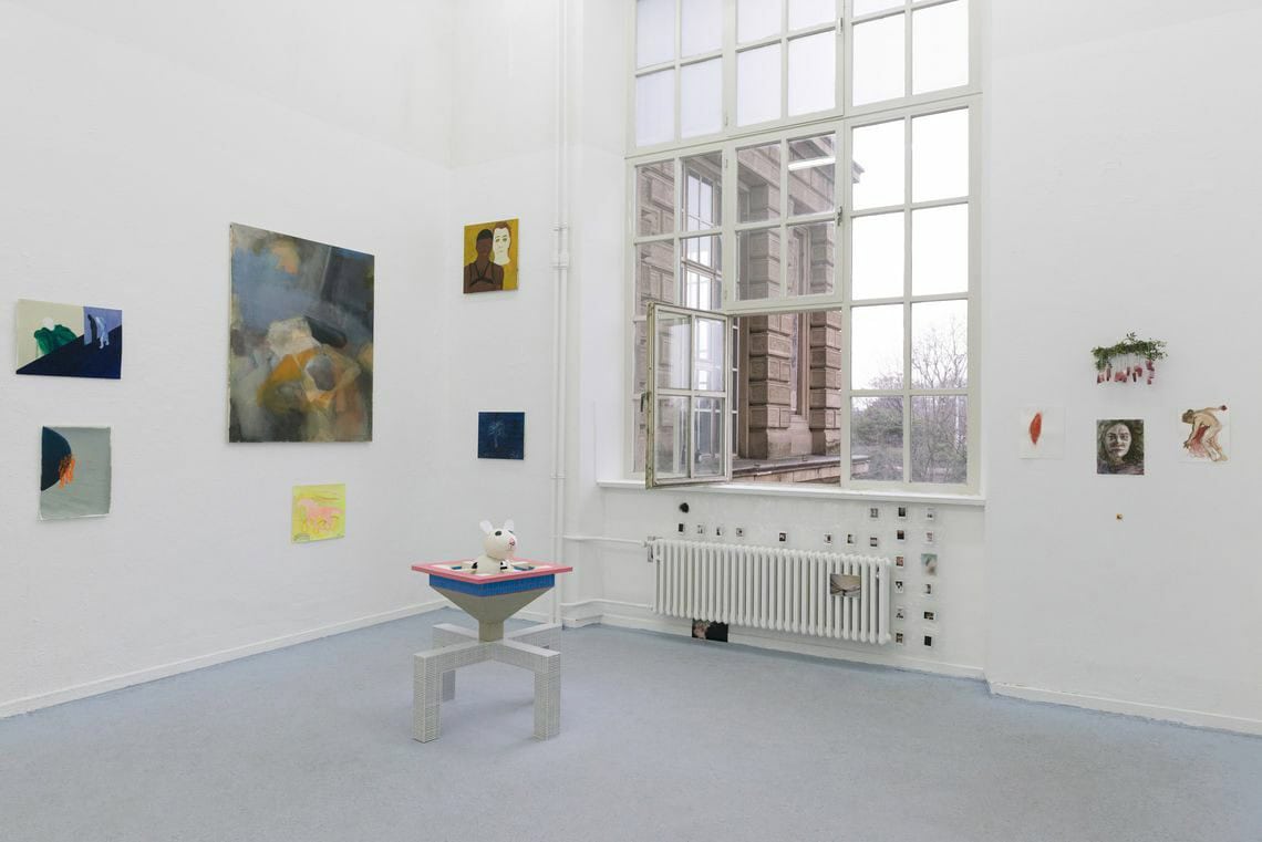 Foto: Ein Raum mit weißen Wänden, grauem Boden und einem großen Fenster. An den Wänden hängen, auf unterschiedlicher Höhe, Arbeiten auf Papier und Leinwand sowie Objekte im Klein- und Mittelformat. Im Raum steht eine Skulptur. Unterhalb der Fensterbank, über, neben und unter der Heizung, hängt ebenfalls eine Gruppe von sehr kleinen Objekten.