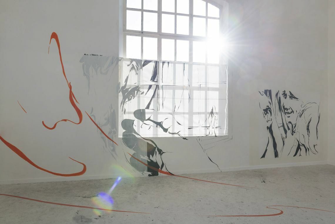 Foto: Ein Raum mit weißen Wänden, hellgrauem Boden und einem großen Fenster. An den Wänden und über dem Fenster hängt ein transparentes Material, auf das schwarze, grafische Elemente gemalt Wurden. Auf die Wand und auf den Boden sind außerdem orangene Linien aufgemalt.