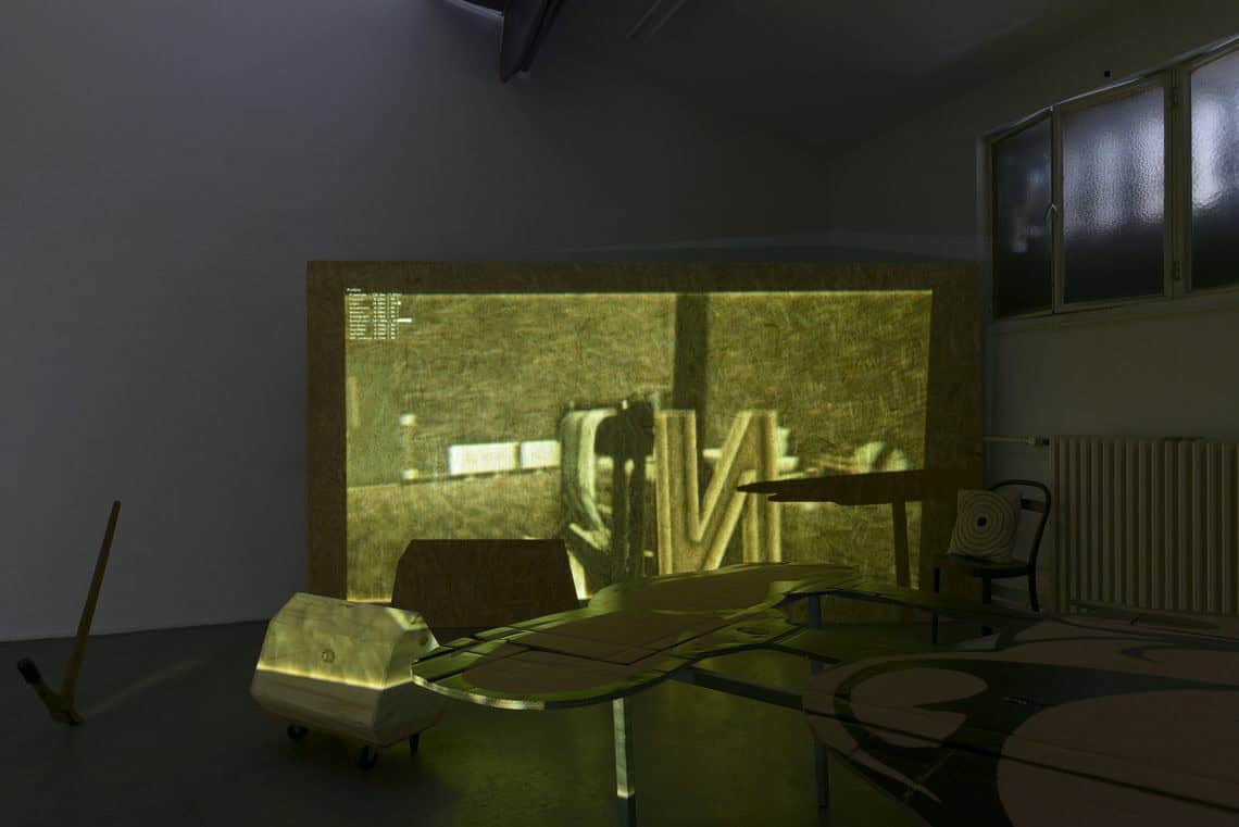 Foto: Ein abgedunkelter Raum. In dem Raum befinden sich mehrere, zum Teil möbelartige Objekte. In der Ecke des Raumes steht eine Platte. Die Platte und Teile der sich im Raum befindenden Objekte werden von einer Projektion angestrahlt.