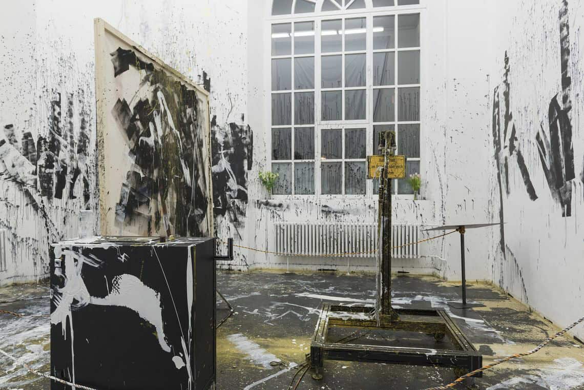 Foto: Ein Raum mit weißen Wänden, dunklem Boden und großem Fenster. Im Raum stehen mehrere Objekte. Die Wände, das Fenster, der Boden und die Objekte sind allesamt mit weißen, schwarzen und gelben Farbsprenklern versehene.