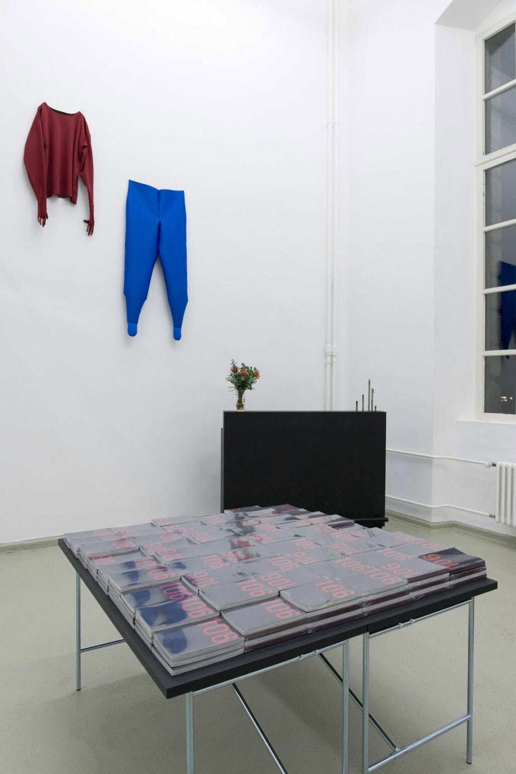 Foto: Die Ecke eines Raumes mit weißen Wänden und grauem Boden. In der Ecke des Raumes steht eine schwarze Verkofferung, auf der ein Blumenstrauß und Objekte stehen. Oben an der Wand hänge zwei kleidungsartige Objekte. Im Raum steht ein Tisch mit schwarzer Tischplatte, der komplett mit gestapelten Heften bedeckt ist, die jeweils in Folie eingeschlagen sind. Auf ihrem Cover steht "106".